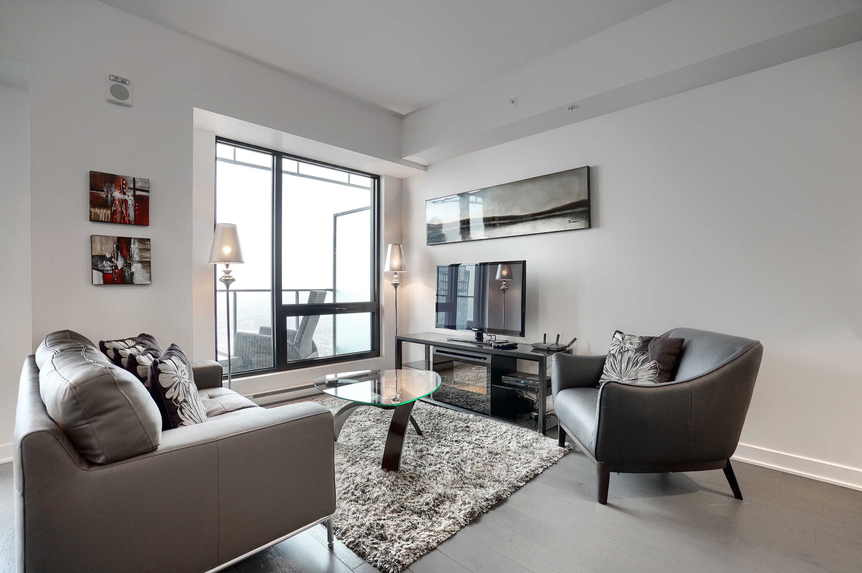 View into the designer living room in this furnished short term housing apartment in montreal. Dark gray leather couch & chair, light fluffy rug under a glass table. Large sunny windows. 