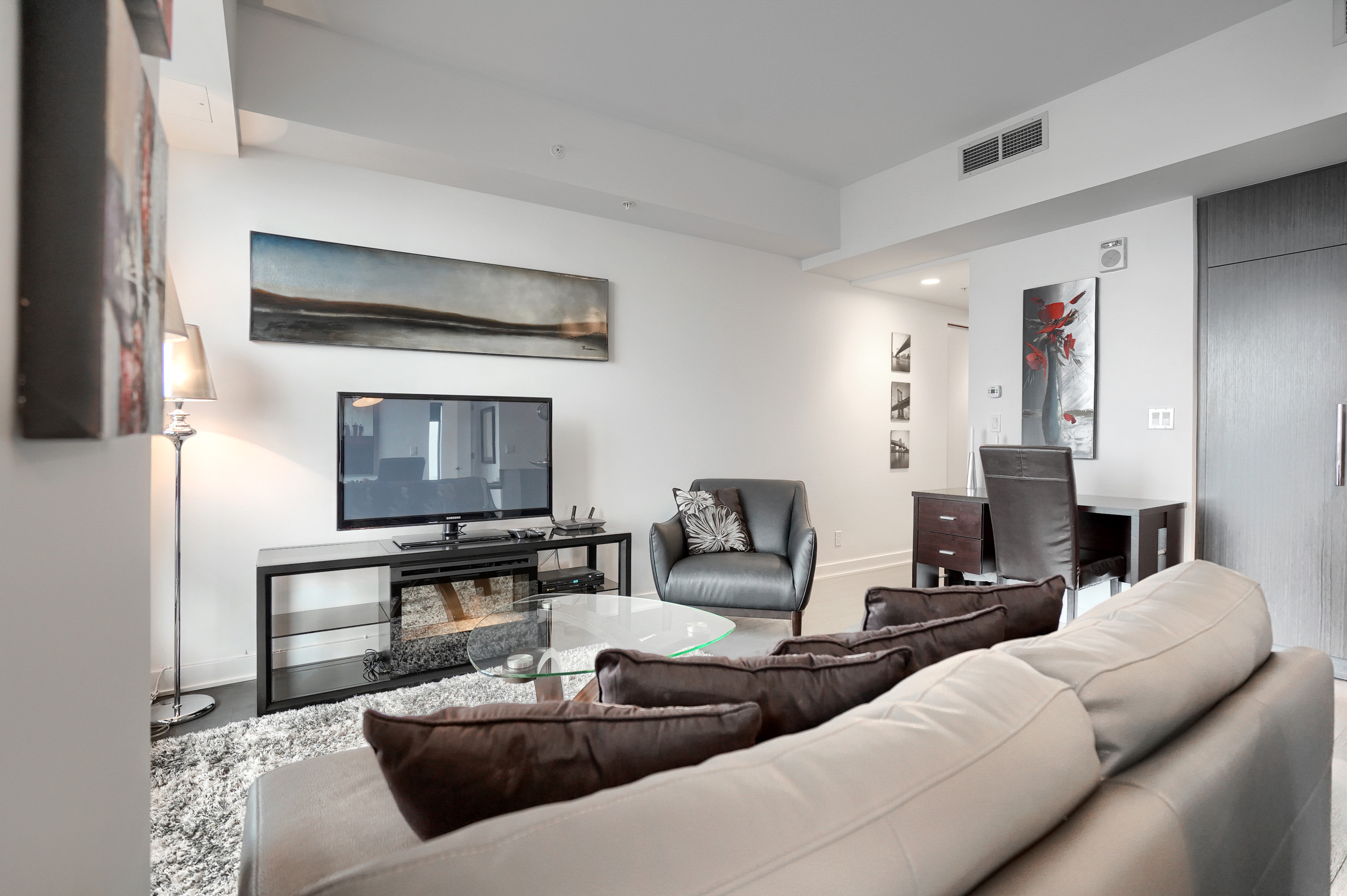 View from the dining area into the living room of this furnished rental in montreal. Gray leather couch & over-sized chair. Flat screen TV, glass table on top of a fluffy rug. Floor-to-ceiling window to the left and study desk with chair to the right. 