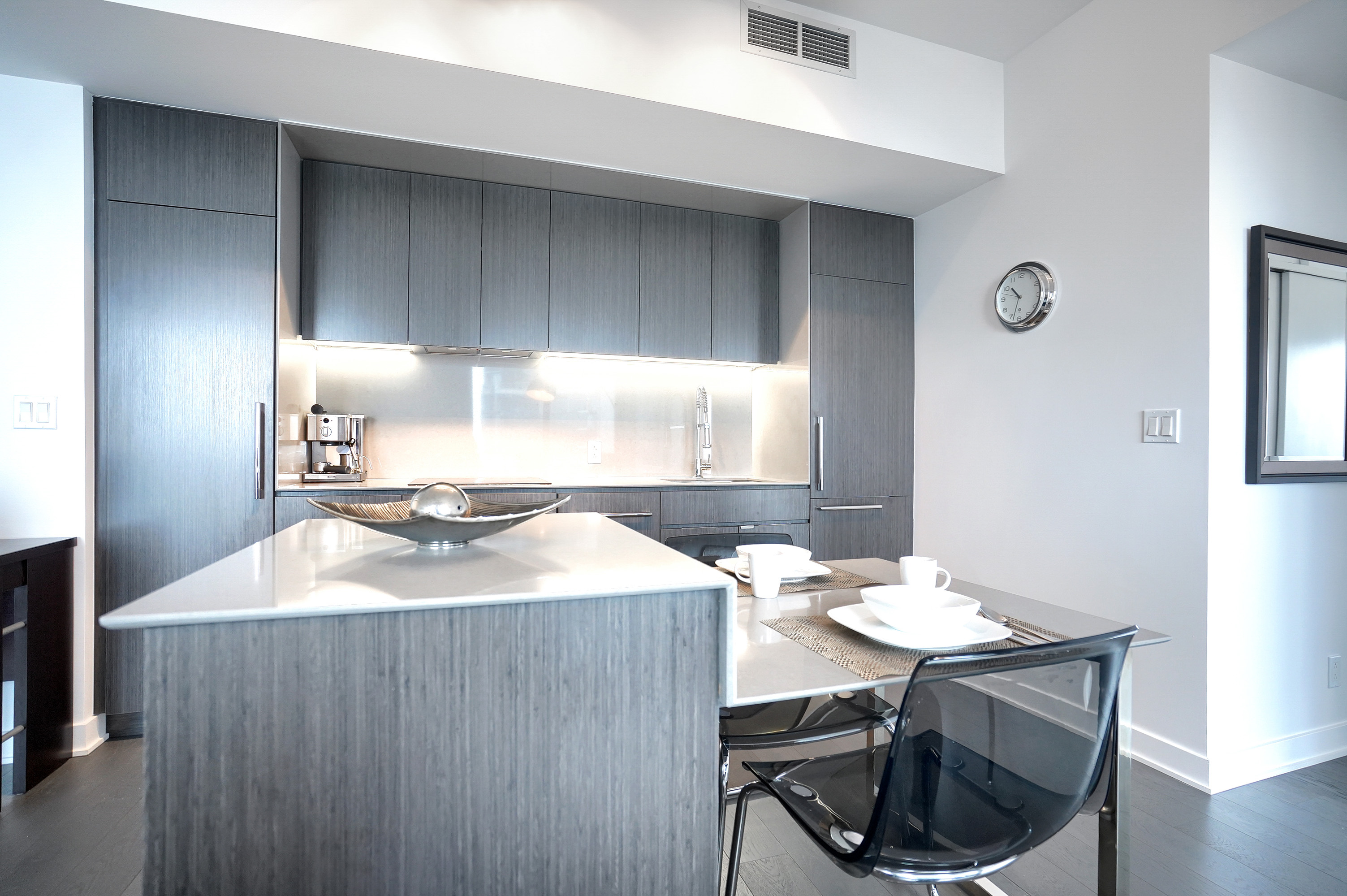 Close-up of the island counter and into the kitchen of this sleek and modern furnished corporate apartment in montreal. Grays and whites. Inset refrigerator and pantry. Flat top stove, overhead lighting and plenty of cabinet space
