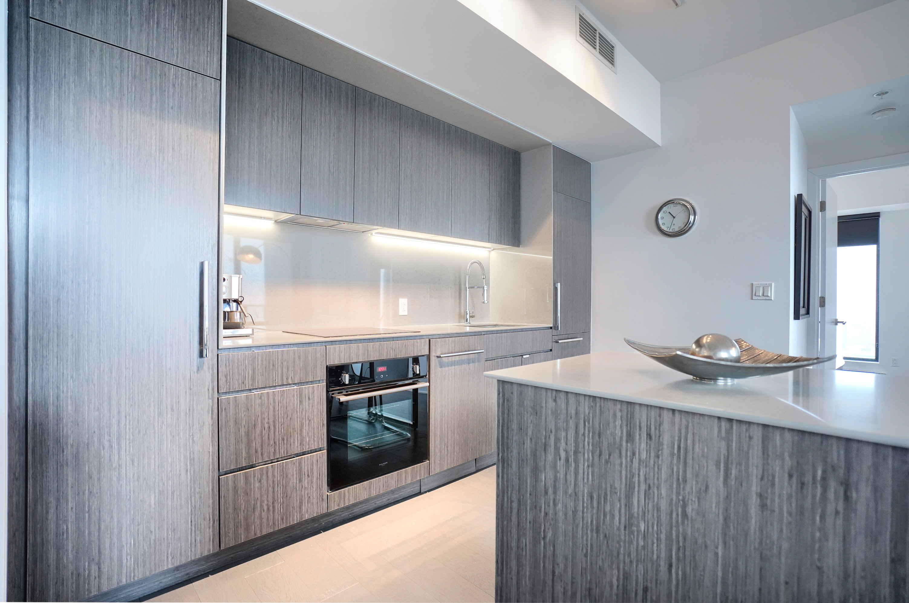 Wide-angle view into the kitchen. Darker gray cabinetry against light gray/white walls. Spacious and modern kitchen with all the amenities in this furnished corporate suite in montreal