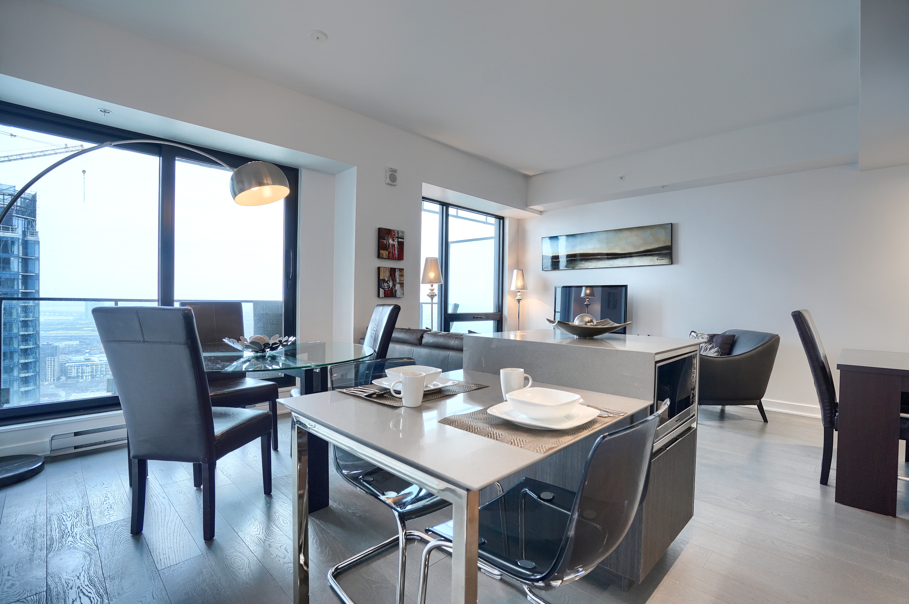 Wide-angle view of the two-seat kitchenette, four-seat glass dining table, plush leather couch, study desk area and two large floor-to-ceiling windows. Luxury everything for your stay in this furnished short term rental in montreal  