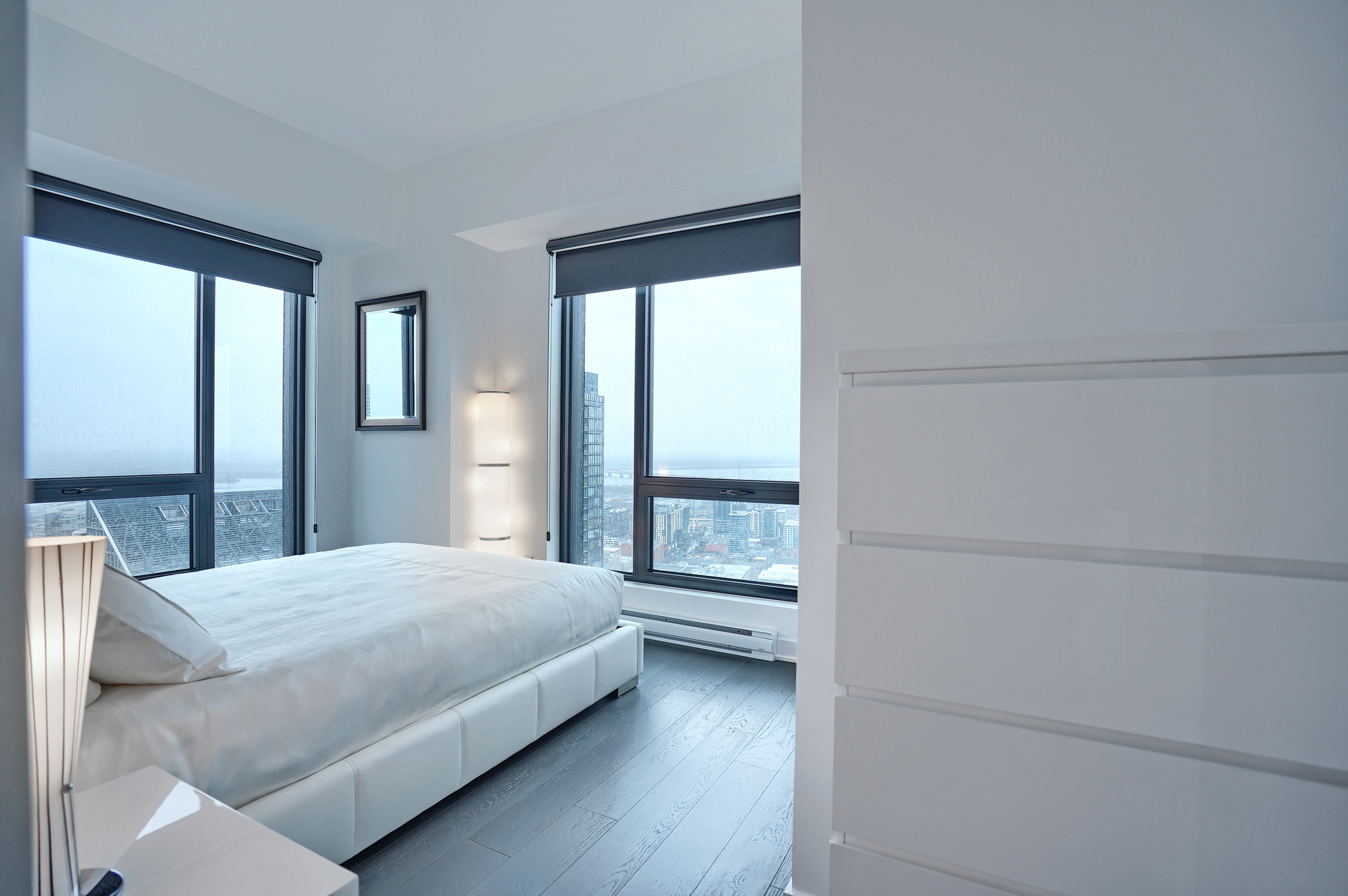 Angled view into the bedroom of this luxury furnished short term housing apartment in montreal. Light, plush bedding, light walls, darker gray wood flooring. Floor-to-ceiling window to brighten each morning