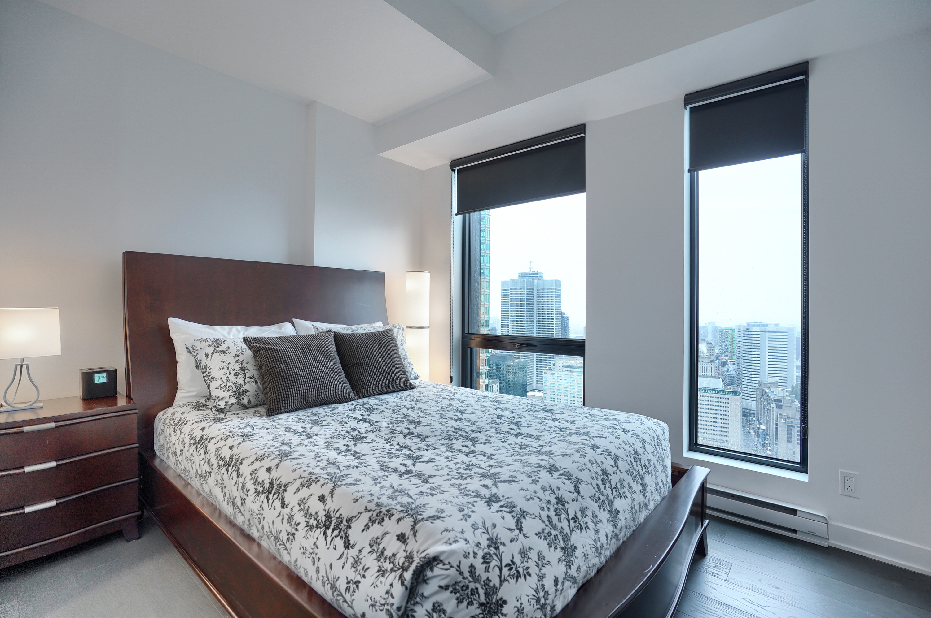 View of the master bedroom in this luxury furnished short term rental in montreal. Comfy white and gray flowered bedding with white and gray accent pillows. Mahogany headboard with matching night tables.  