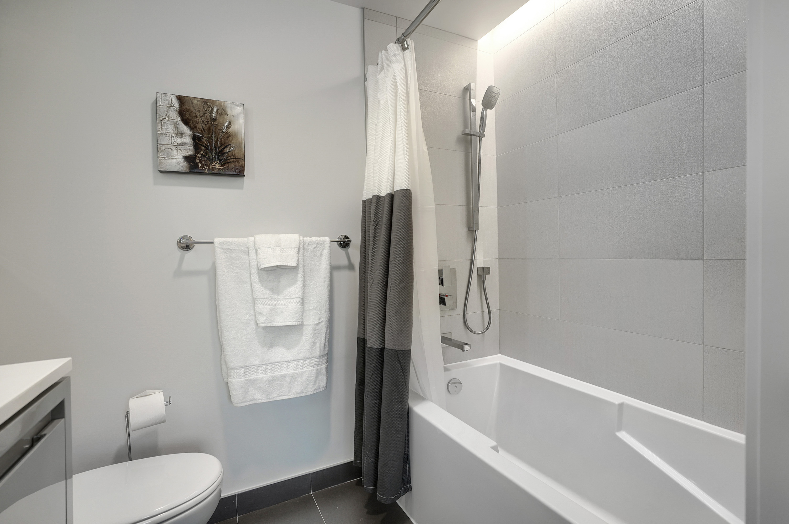 Angled view of the modern bathroom in this luxury furnished short term housing apartment in montreal. Modern white tube with squared edges, adjustable shower head, white plush towels. White and gray accents throughout 