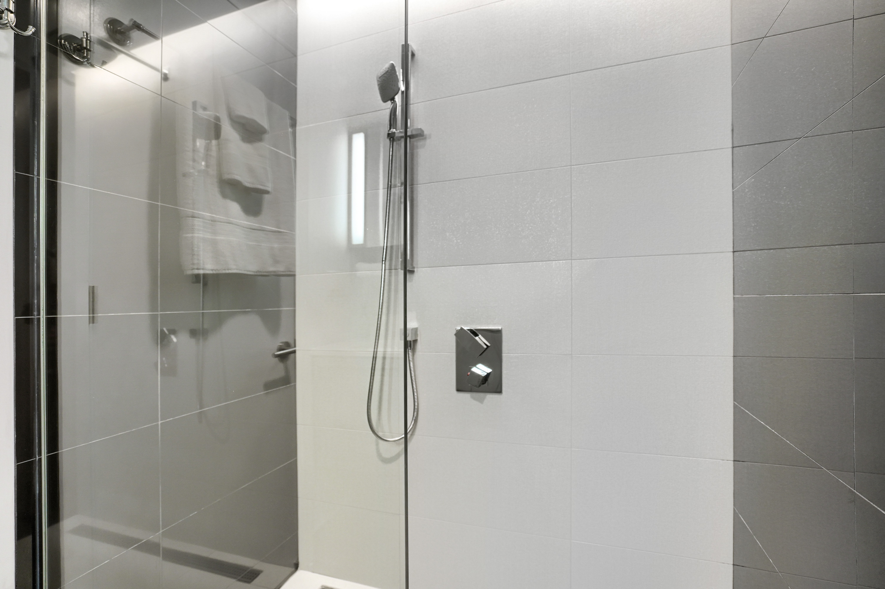 Close-up of the floor-to-ceiling glass shower with adjustable stainless shower head in the master bath of this furnished executive housing apartment in montreal. Stainless accents, white and gray sleek tile.