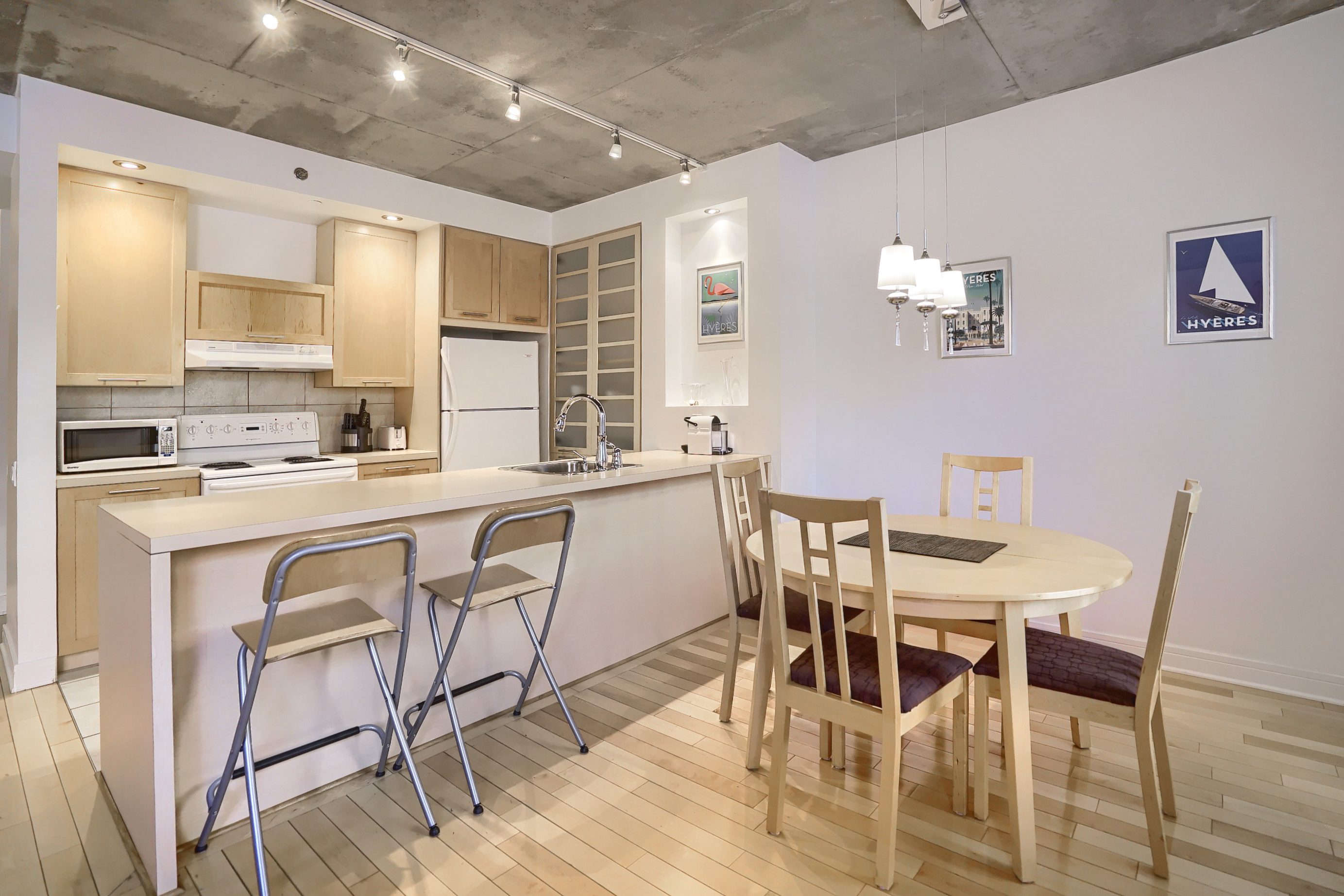 View of the dining table and four wooden chairs, unique stainless-steel ceiling lighting, white walls with light wood floor. Our goal is to provide corporate housing in Montreal that exceeds all your expectations