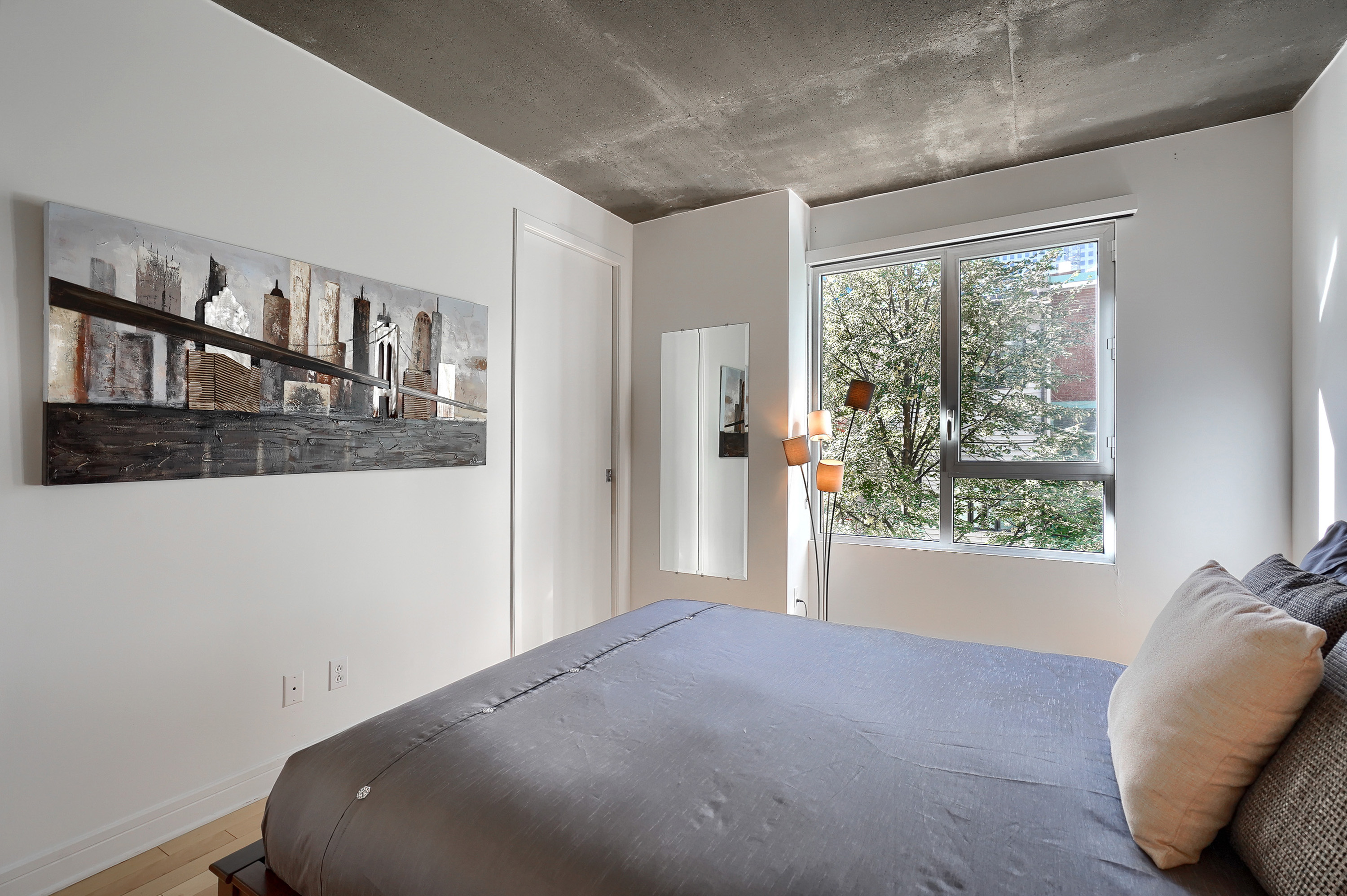 Vue du pied de lit avec tableau moderne dans les tons de brun et argent sur mur blanc, long miroir sur la droite et fenêtre beignant la chambre de soleil tout au long de la journée dans cet appartement entièrement équipé au centre-ville de Montreal