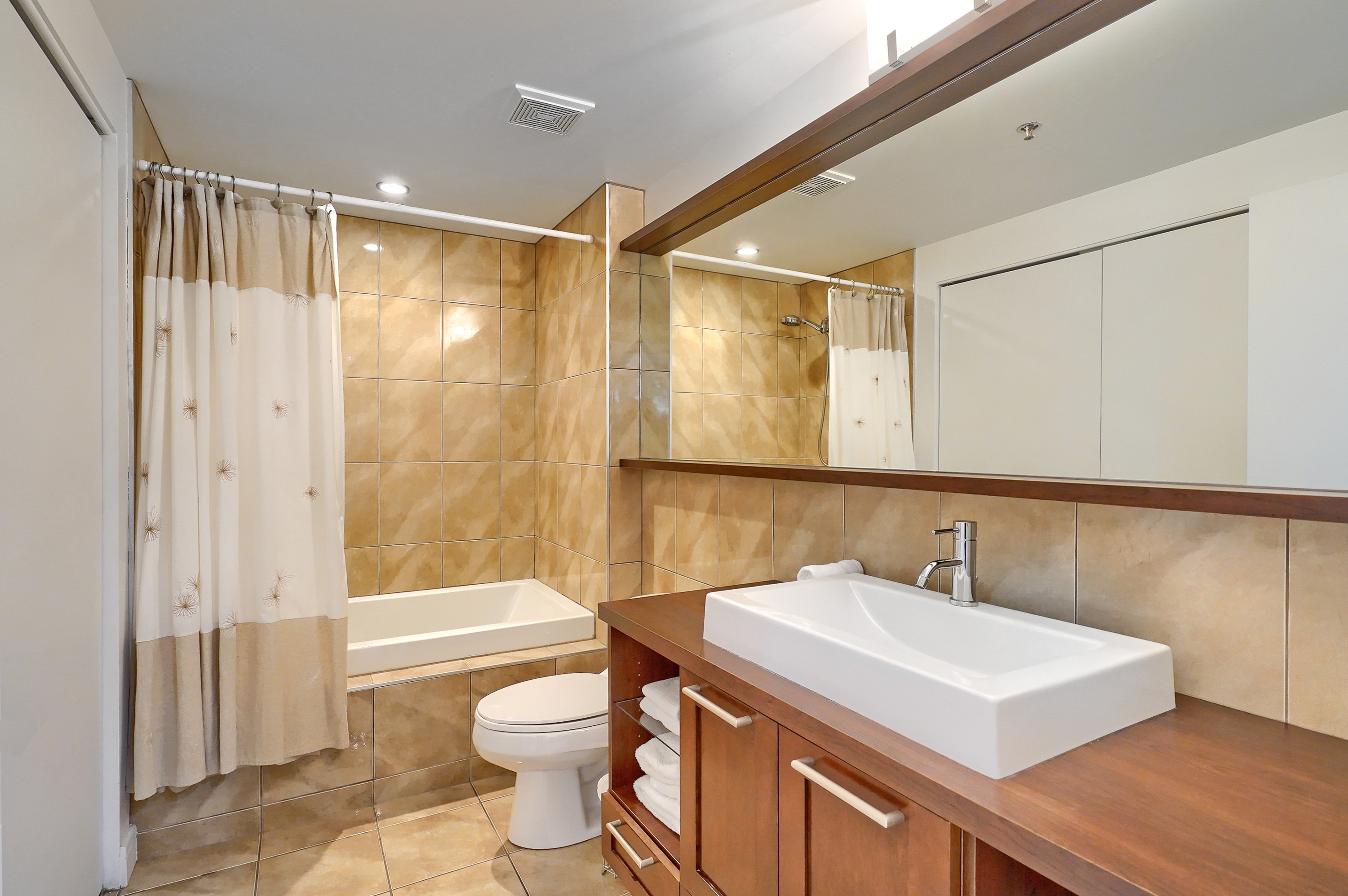 Vue en angle de la salle de bain montrant la toilette et le lavabo carré unique, moderne, blanc et épuré. Aussi, nous voyons la baignoire spacieuse et le miroir et l'éclairage dans cette salle de bain moderne. Un avantage de ce logement meublé à Montréal 