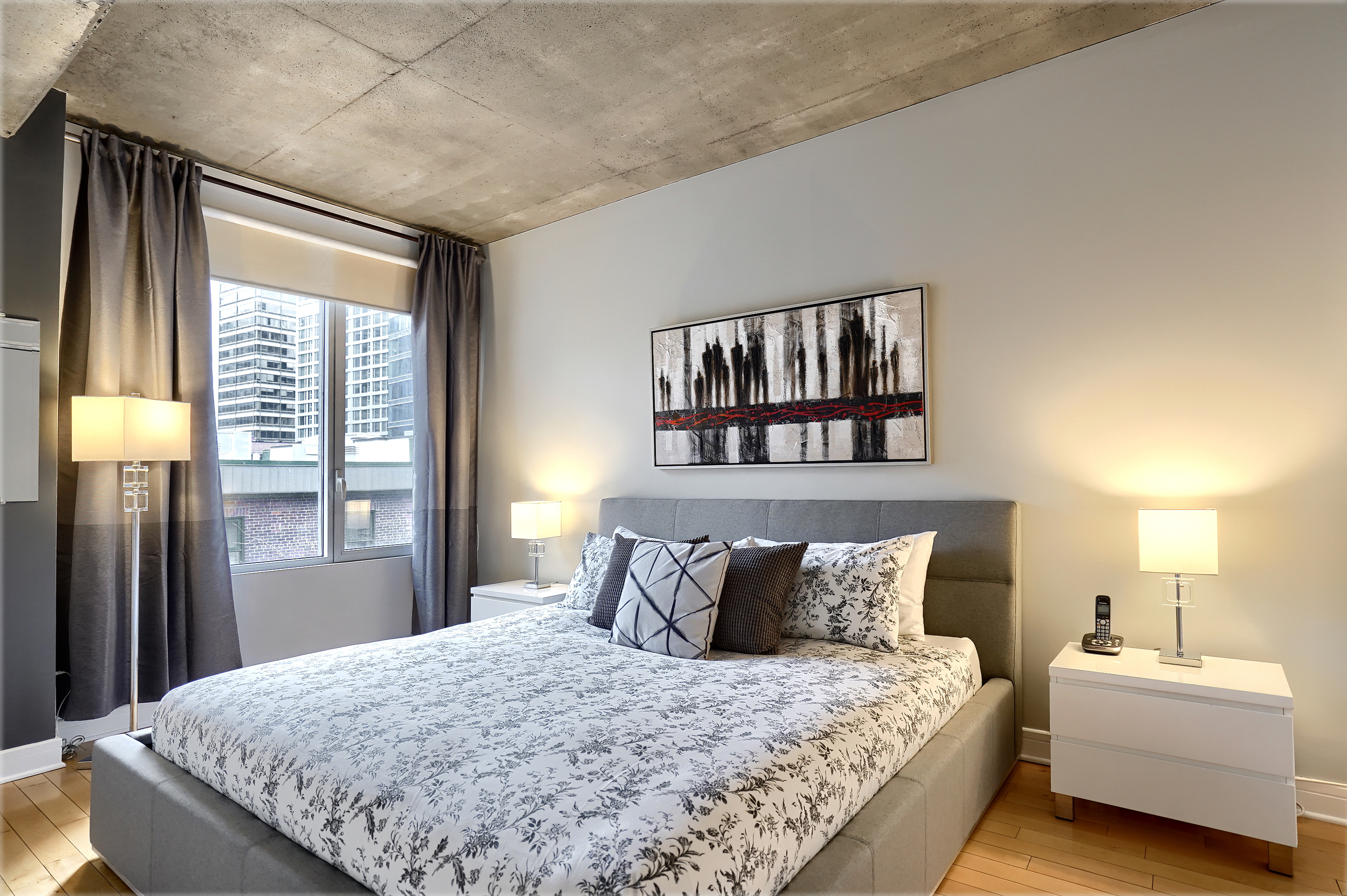Wide-angled view of the bedroom showing the king size bed and its white and grey comforter and accent pillows and cushions, light grey headboard and frame, white night tables with a lamps, a large window of this furnished short-term rental in Montreal