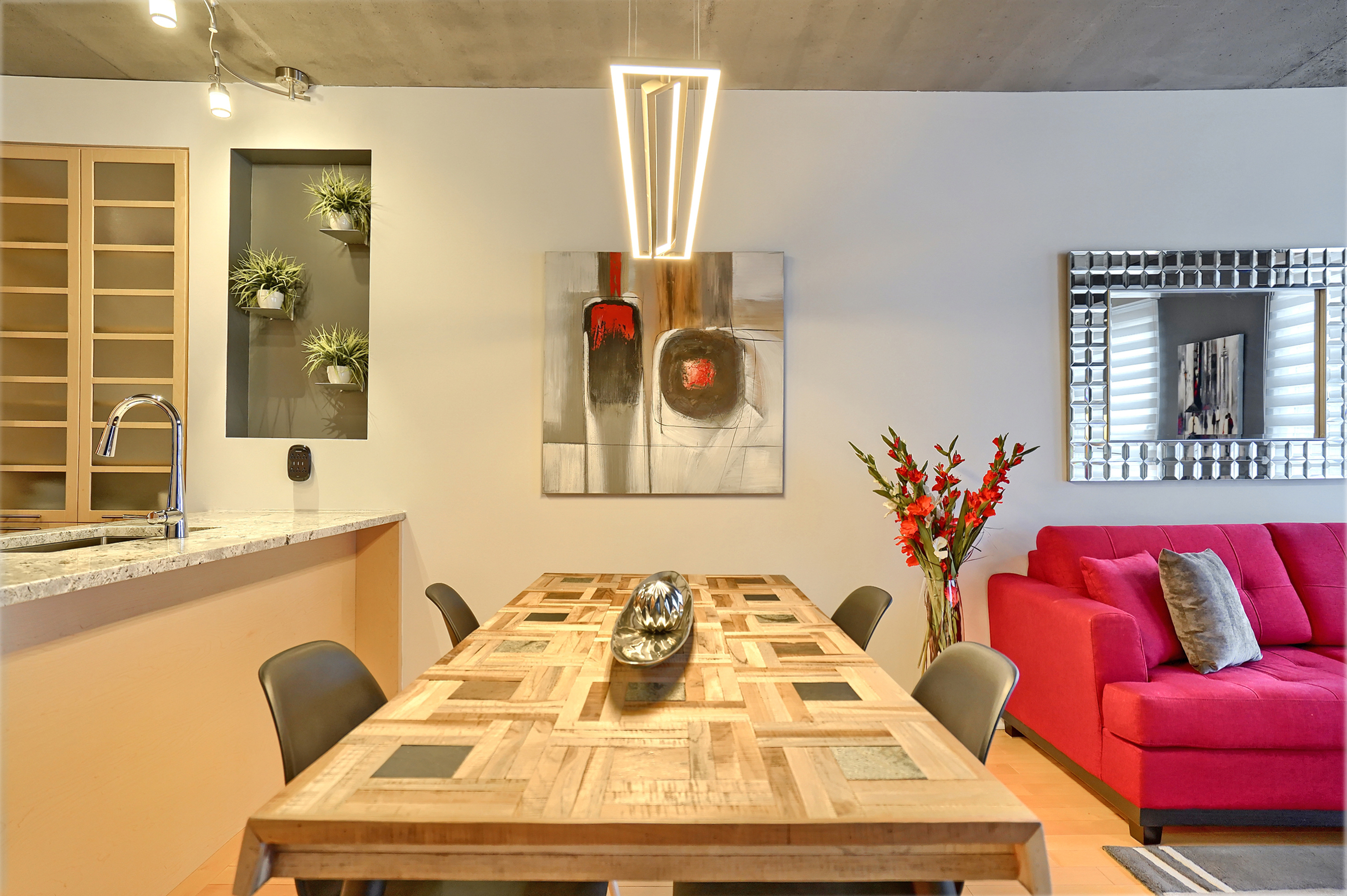 View of the designed wooded dining table and a modern DEL fixture and a painting with red colors matching perfectly with the red sofa on the right in this cozy 
apartment de dedicated to corporate housing in Montreal