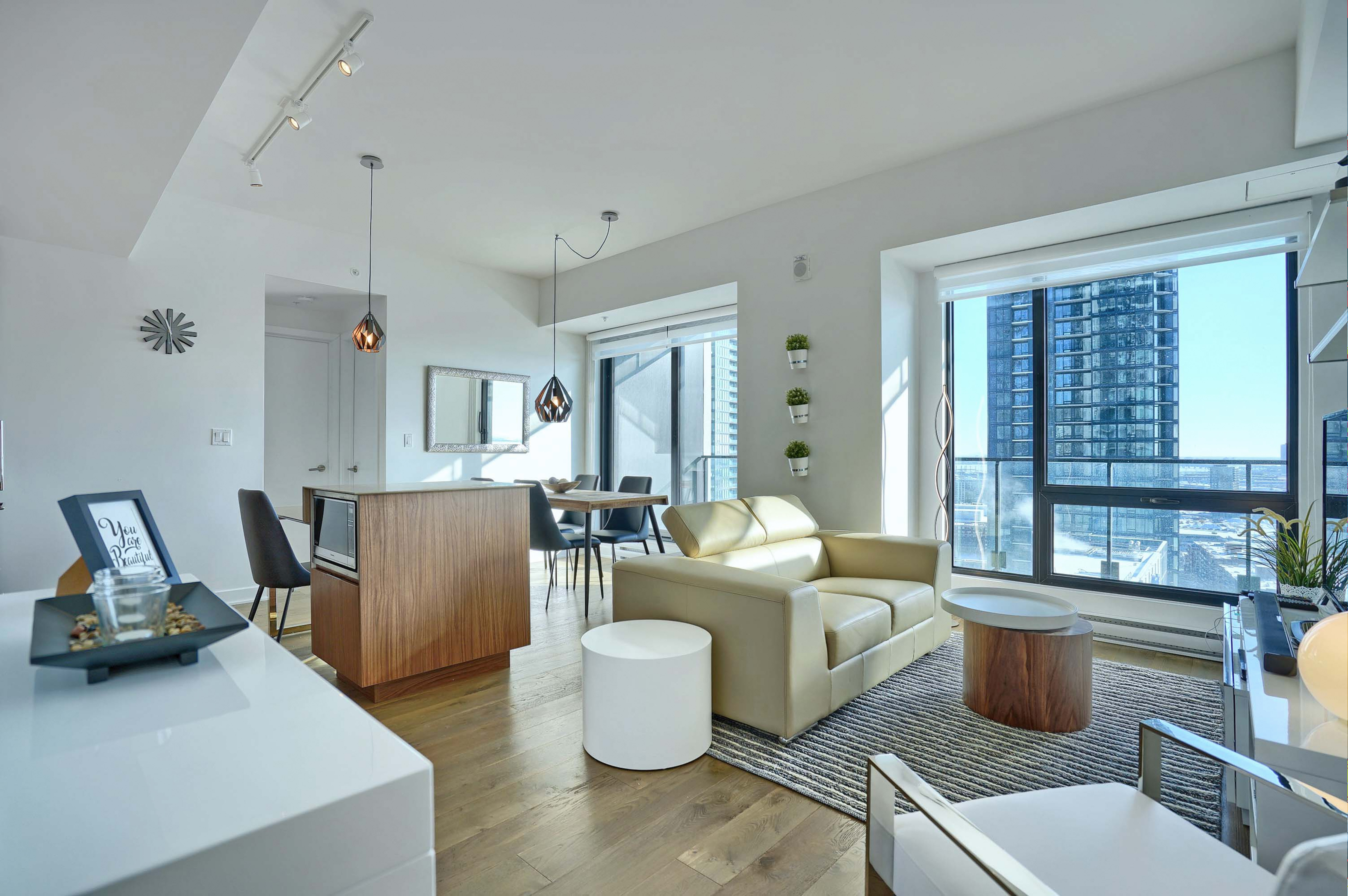 View of the main living and dining areas of this furnished suite in montreal. Luxury, designer furnishing include a leather couch, unique end tables, dining seating for four with light wood flooring, and white walls. Large floor-to-ceiling windows make this an amazing place to be while in montreal. 
