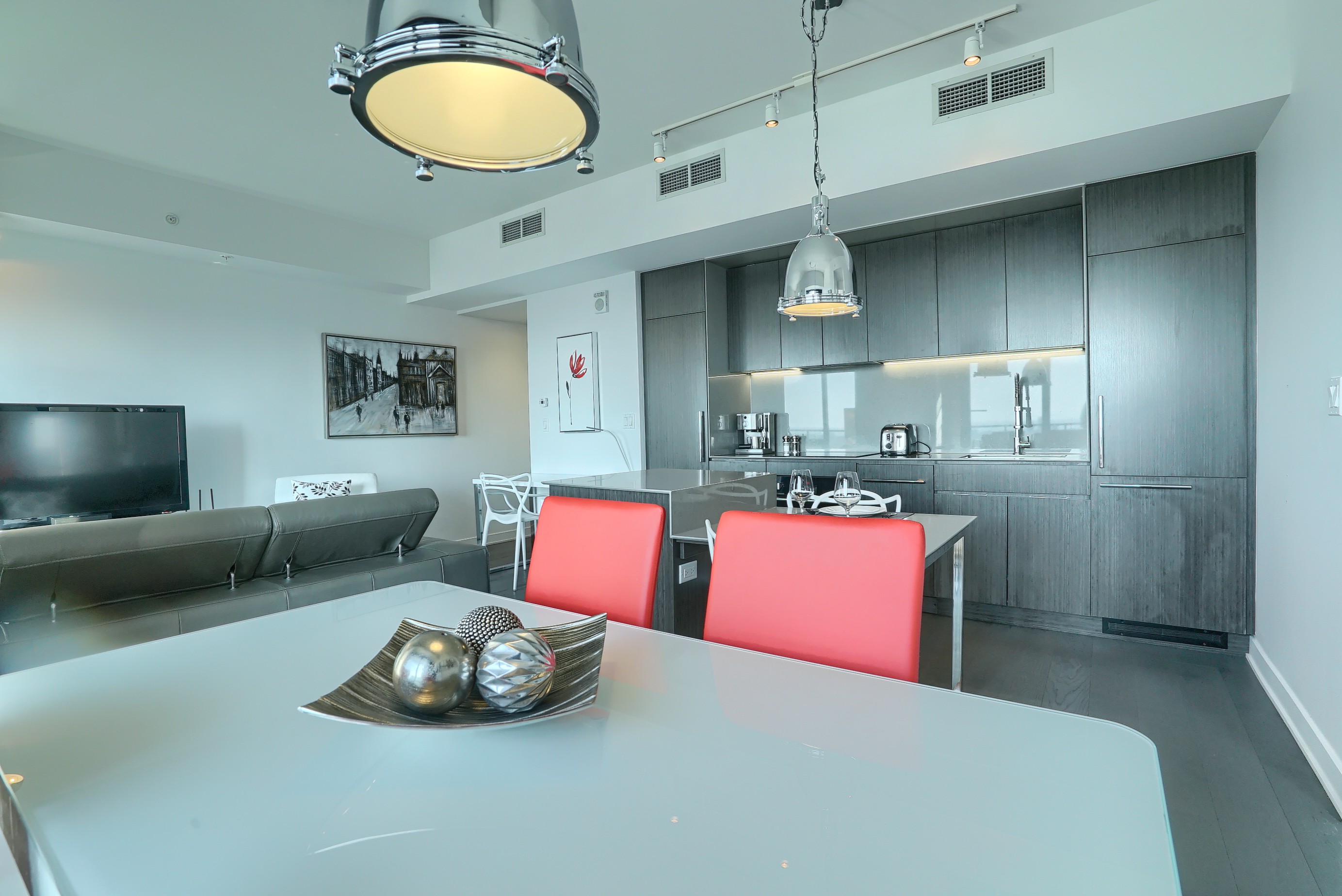 Gros plan de la table blanche à manger moderne avec des chaises rouges. Vous pouvez également voir dans la cuisine et le salon avec un canapé en cuir et une télévision grand écran dans cet appartement entièrement meublé à louer à Montréal 
