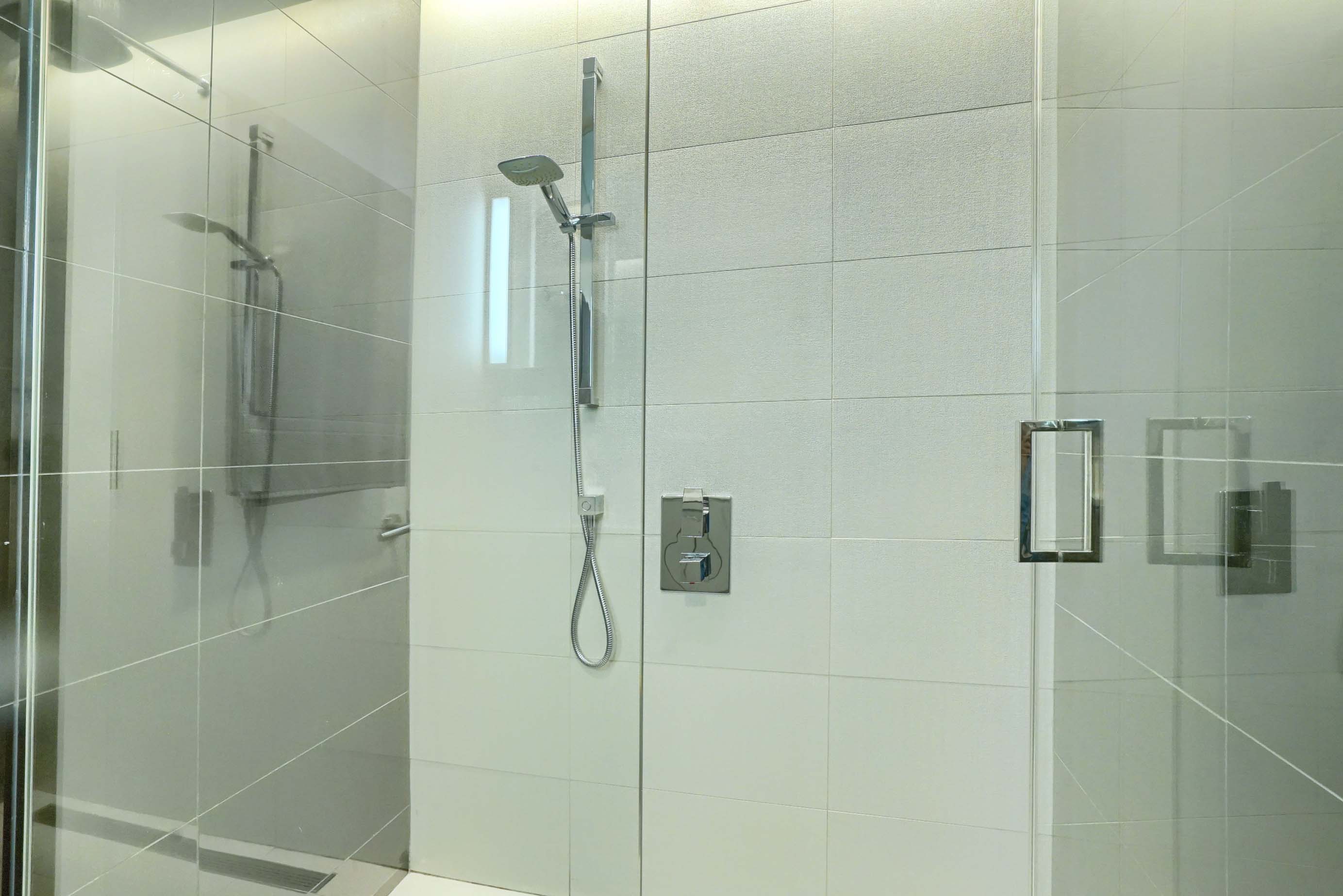 Vue élargie de la douche en verre du sol au plafond avec des robinets modernes en acier inoxydable, des poignées et une pomme de douche réglable. Une salle de bain parfaite pour cet appartement de luxe meublé de luxe à Montréal
