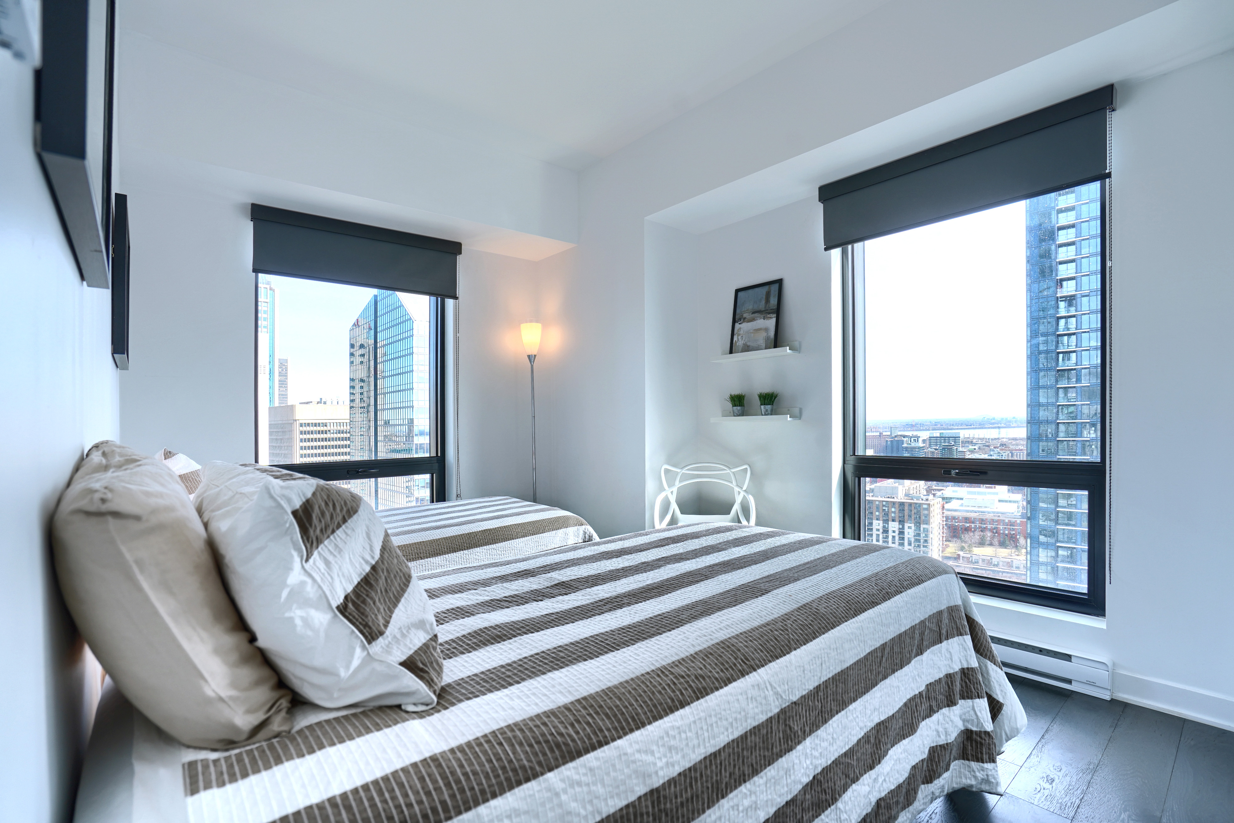 Angled view of the second bedroom showing both single beds, designer plush bedding, two floor-to-ceiling windows brining in sunshine throughout this furnished short term rental in montreal