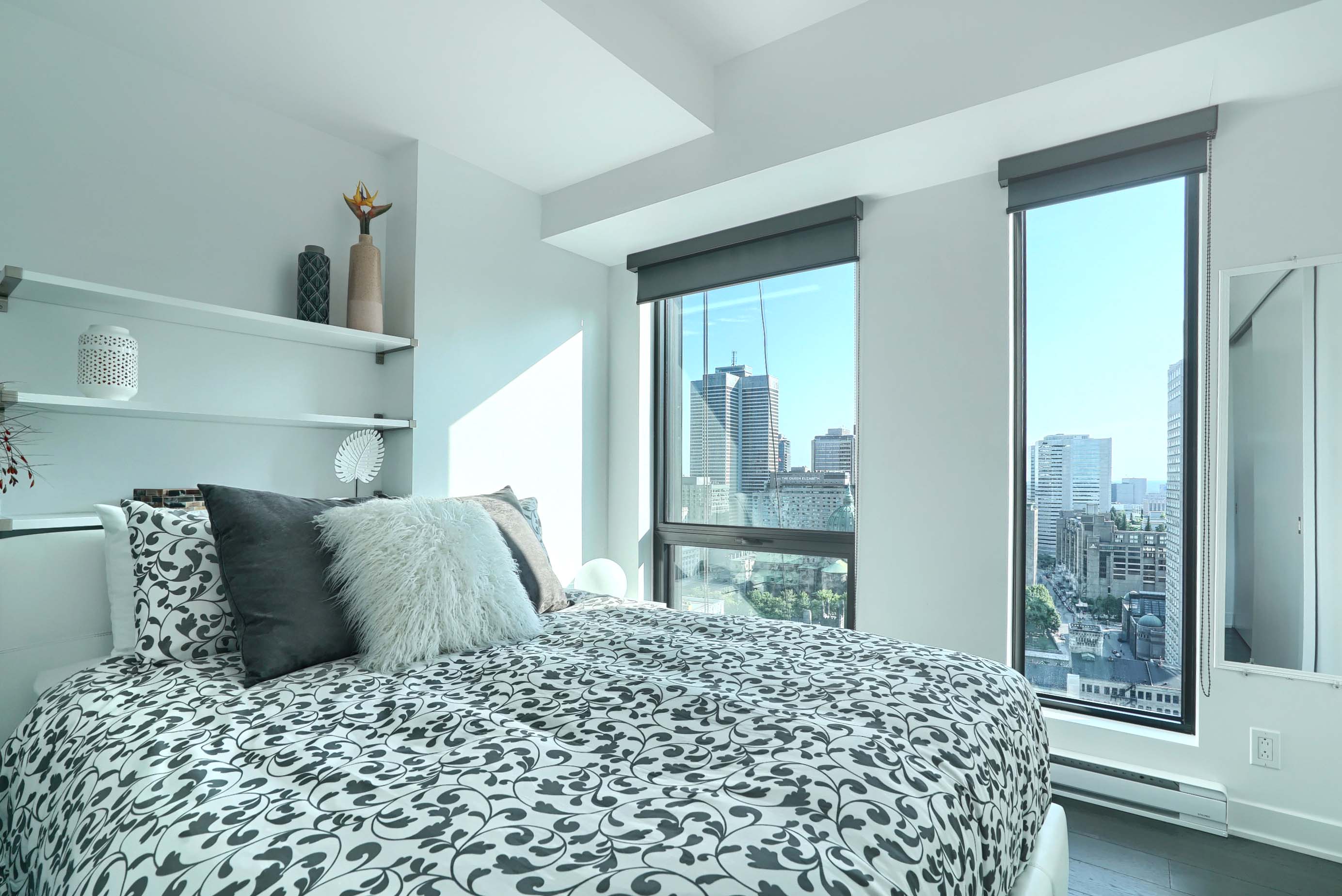 Angled view of the bedroom showing the white and gray plush comforter, fluffy accent pillows, designer shelving and floor-to-ceiling window in this spécialiste en appartements meublés corporatifs à Montréal 