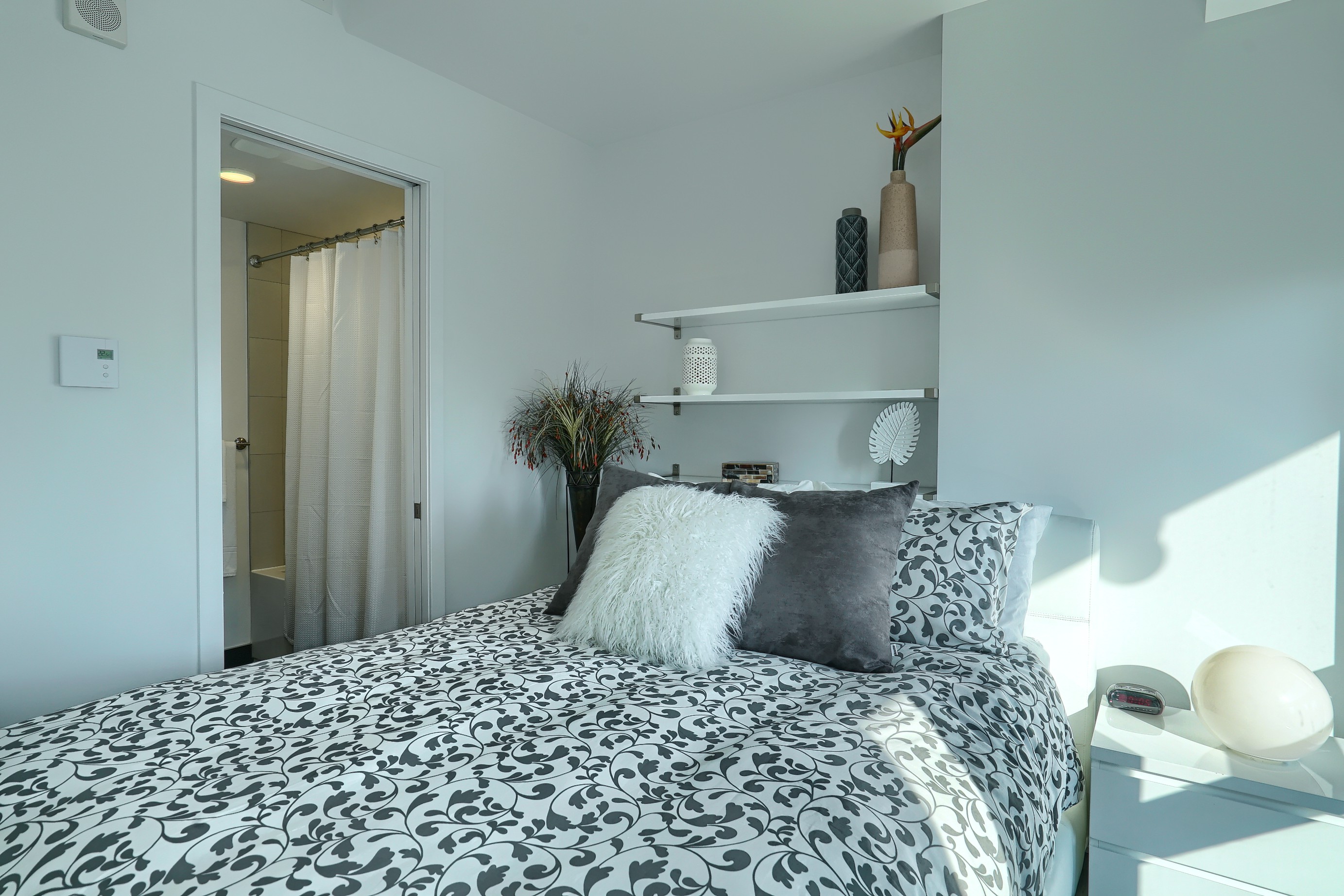 Uniquely designed bedroom with queen-sized bed, plush bedding with white and gray fluffy pillows to accent. White shelving and white drawers enhance this luxury furnished temporary housing apartment in Montreal