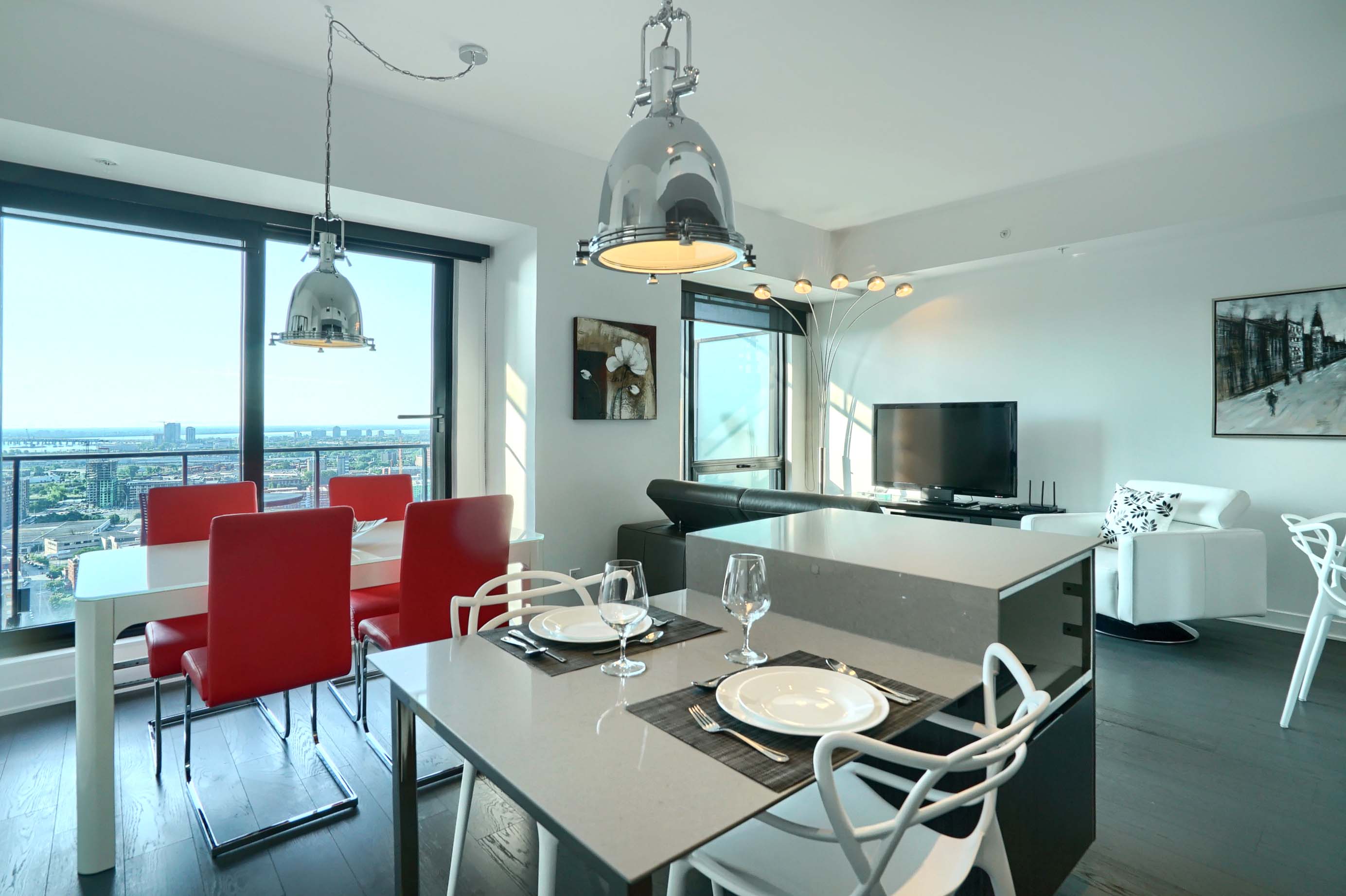 View of the living space of this furnished rental in montreal. Dining table with seating for four, kitchenette with seating for two, living room, flat screen TV and floor-to-ceiling windows for a sunny stay in montreal