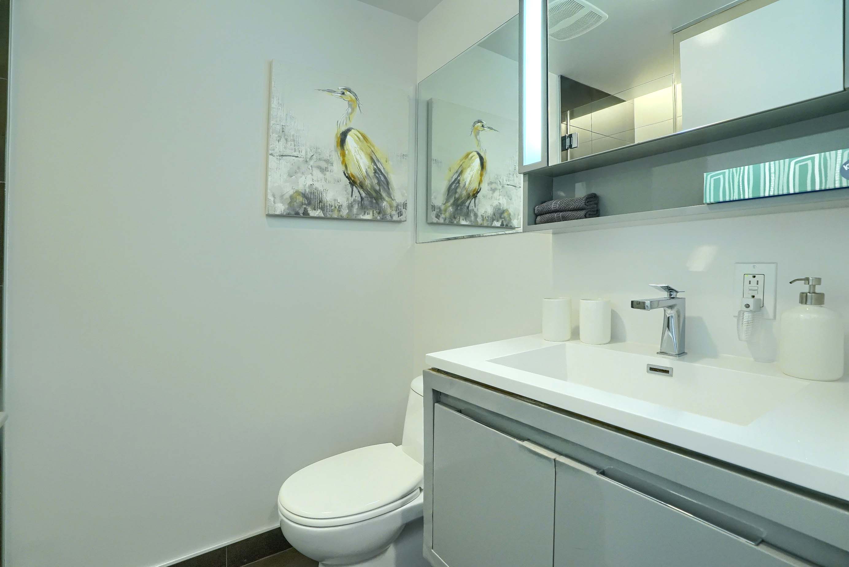 View of the bathroom showing the oversized white sink with designer faucet, spacious white counter, toilet and mirrors all around in this luxury furnished rental, short term, in montreal