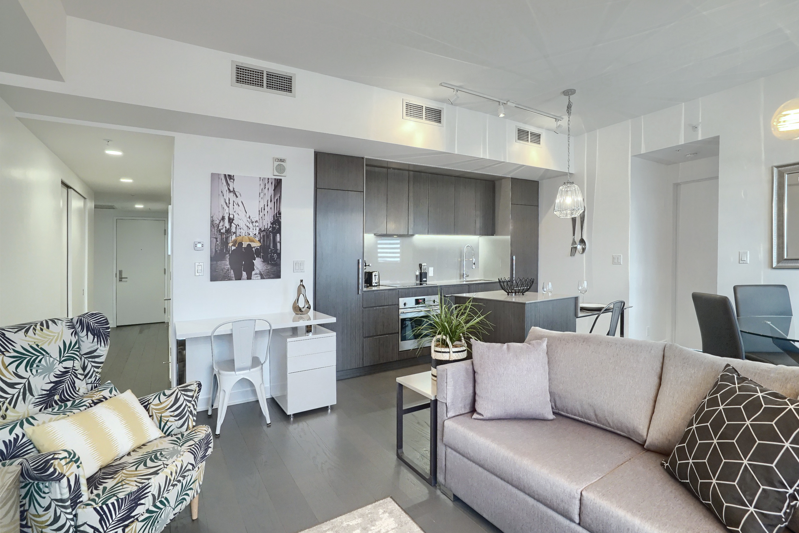 View into the living room and kitchen areas. Light gray cloth couch, white walls, darker wood flooring, decor accents throughout. Desk with chair and modern kitchen make this furnished rental in montreal perfect