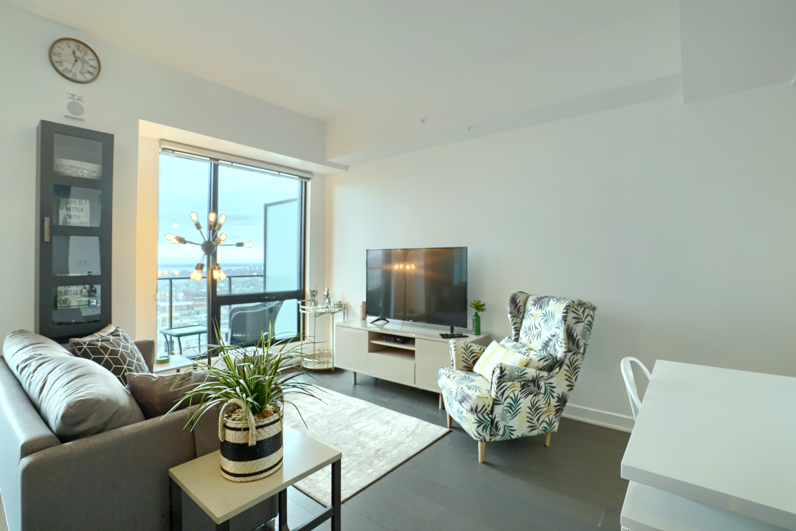 Side view of the living area showing the floor-to-ceiling window, designer couch and chair, flat screen TV, white walls and darker wood flooring. A perfect furnished corporate apartment in montreal