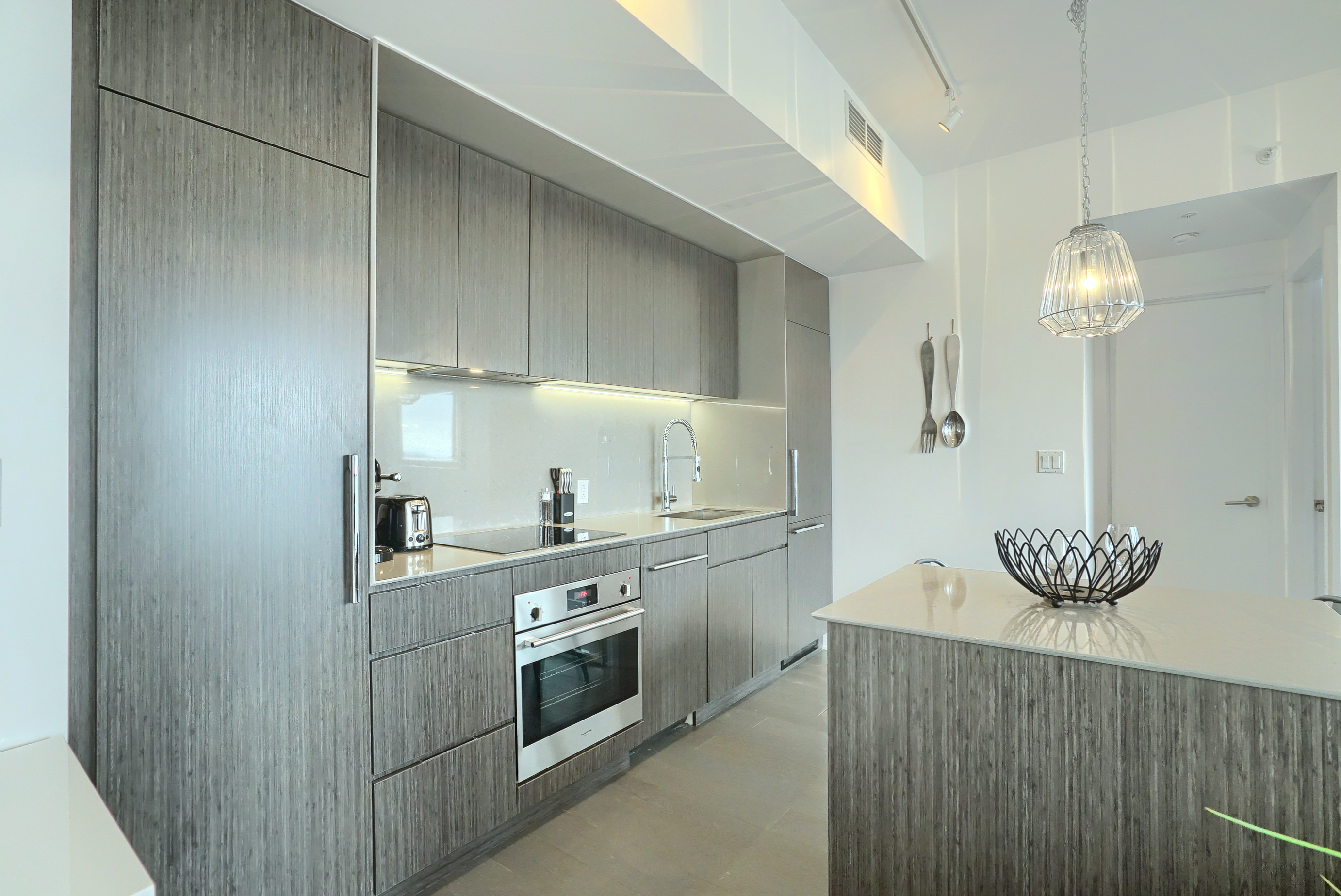 Angled view into the modern kitchen of this furnished rental in montreal. Darker gray cabinets with inset pantry and refrigerator. Generous counter space, flat top stove and inset oven. Light and spacious, ultra modern kitchen 