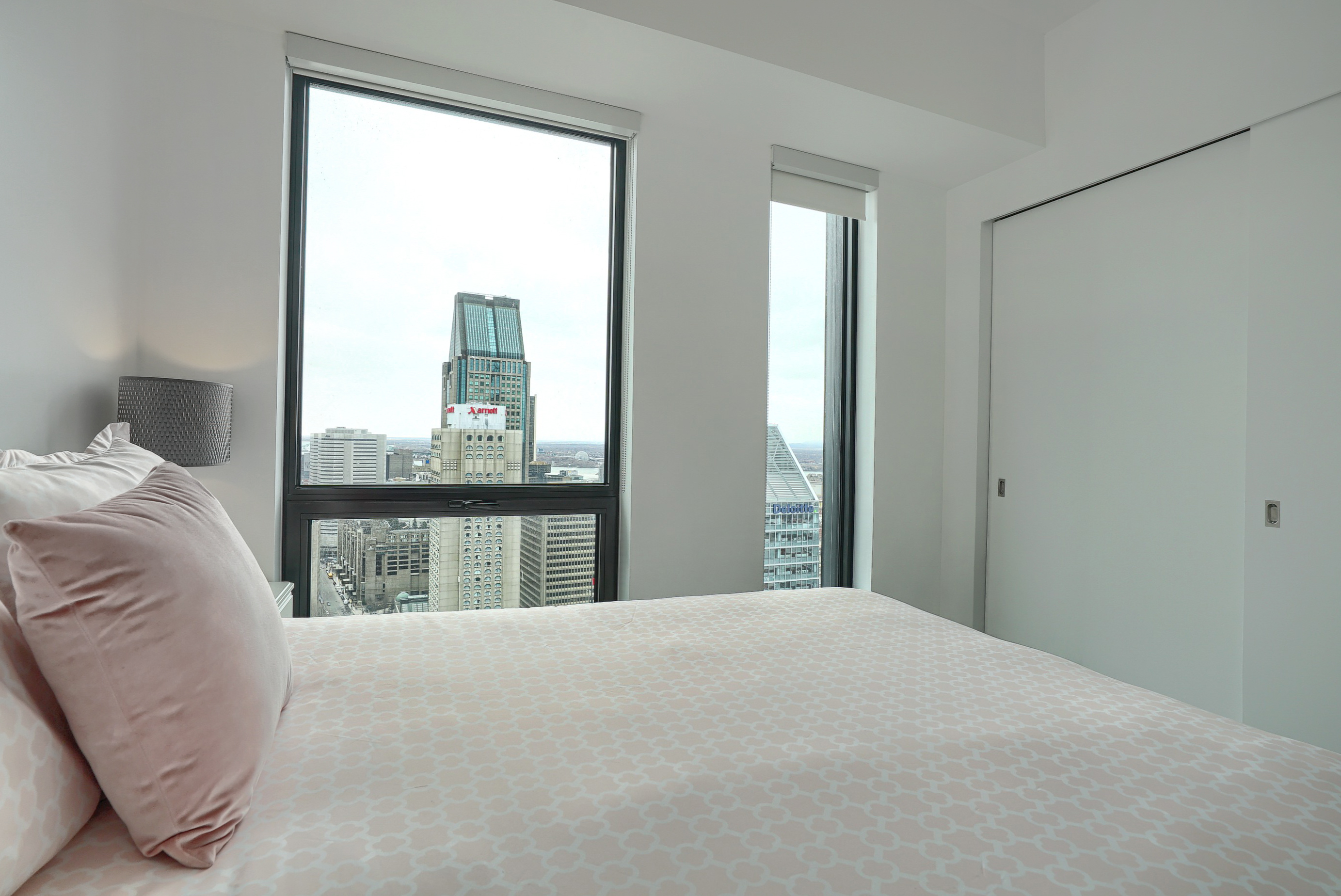 Second bedroom showing the plush light pink and white bedding with designer pillow accents. Large walk in closet with sliding doors and two floor-to-ceiling windows for plenty of light in this furnished corporate housing apartment in montreal