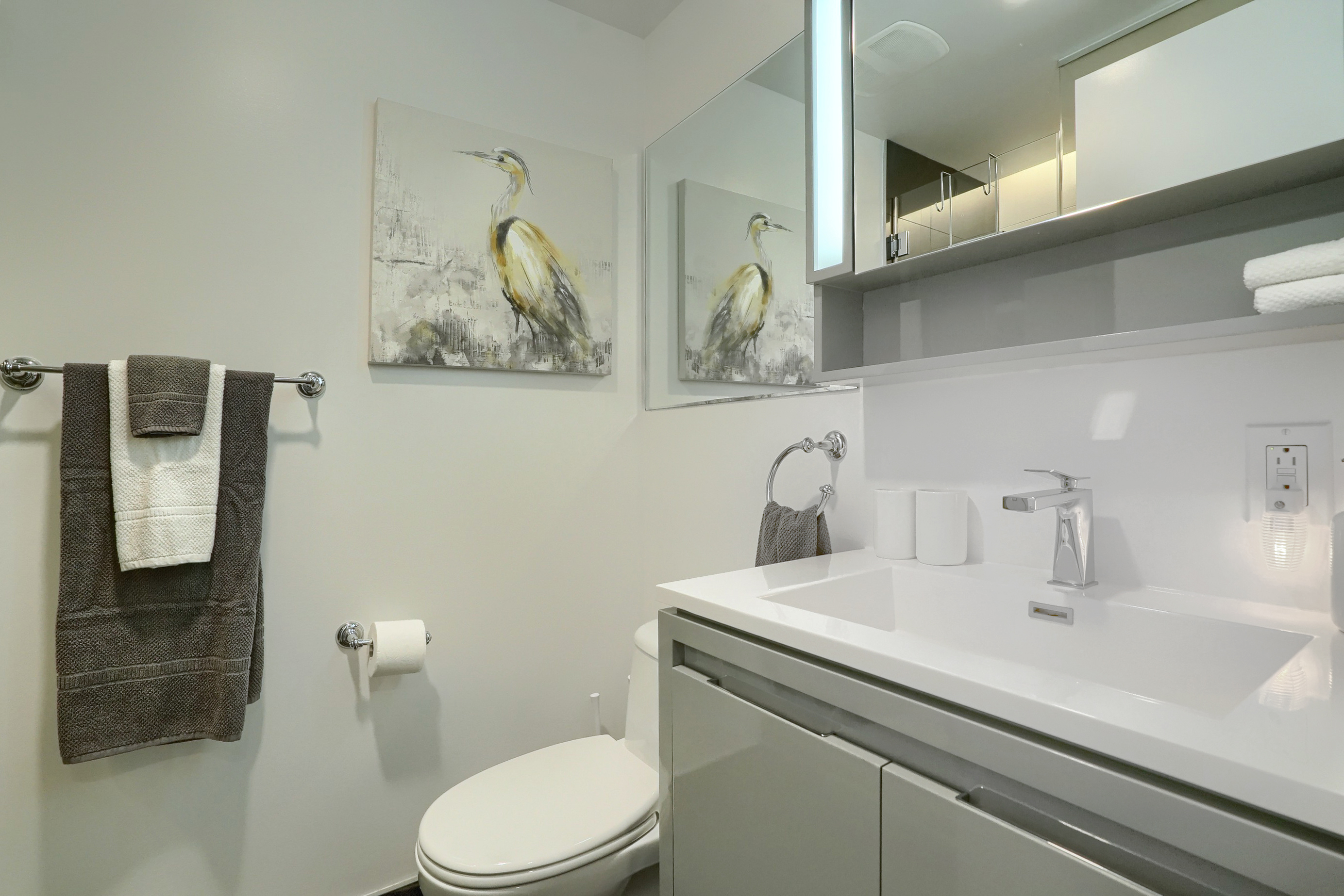 Angled view into the modern and sleek bathroom. Whites and grays. Over-sized white sink with designer stainless faucet. Plush white and gray towels to enhance. Spacious bathroom in this luxury furnished executive housing apartment in montreal