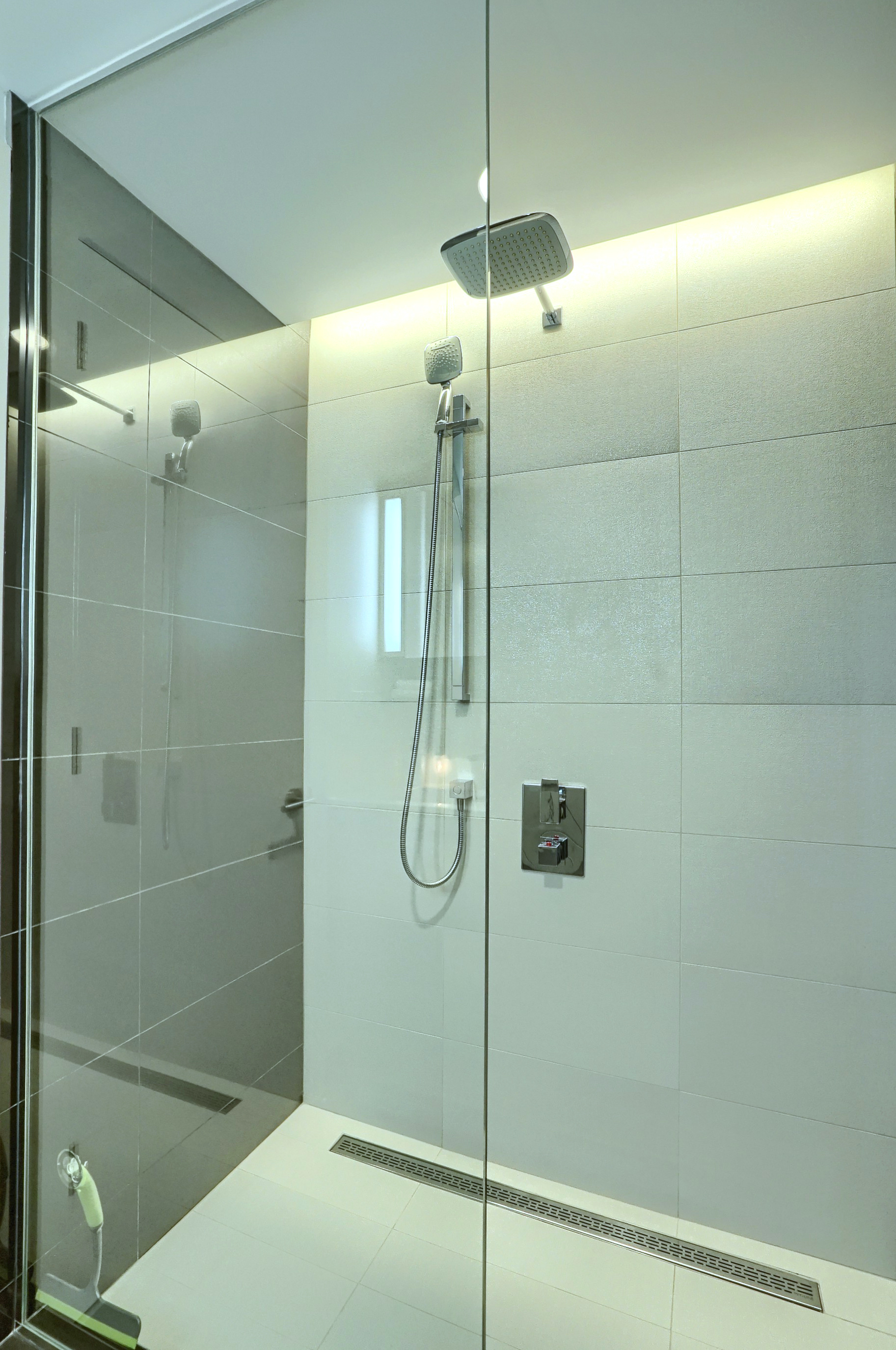 Angled view of the floor-to-ceiling glass shower with adjustable shower head. For cleaning convenience, there is a designer glass cleaning tool. White and gray sleek tile. Spacious modern bathroom in this furnished rental, short term, in montreal