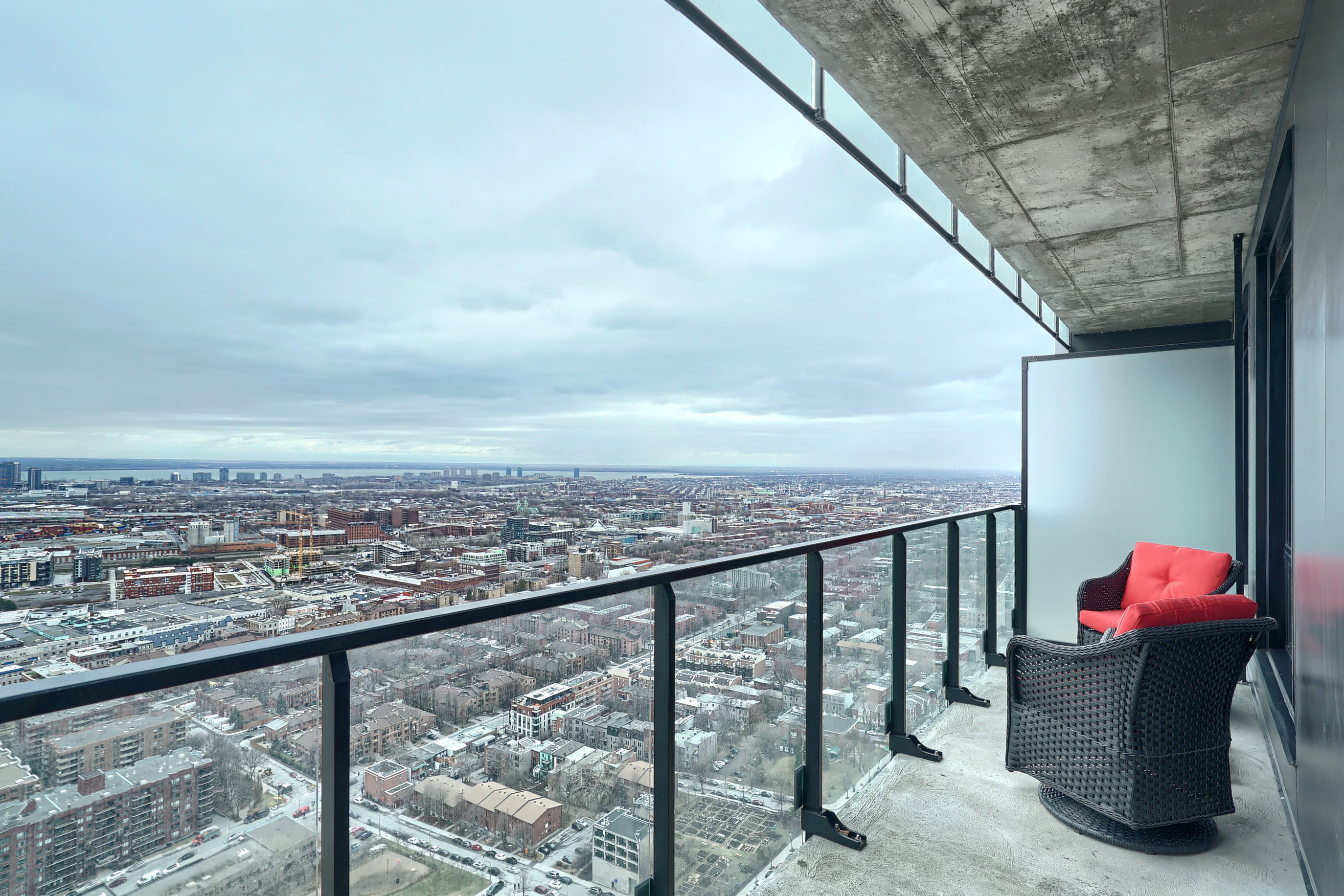 Side view of the balcony showing the glass railing, two designer gray chairs with red accent pillows and a wonderful view of this city from this short term stay apartment in montreal