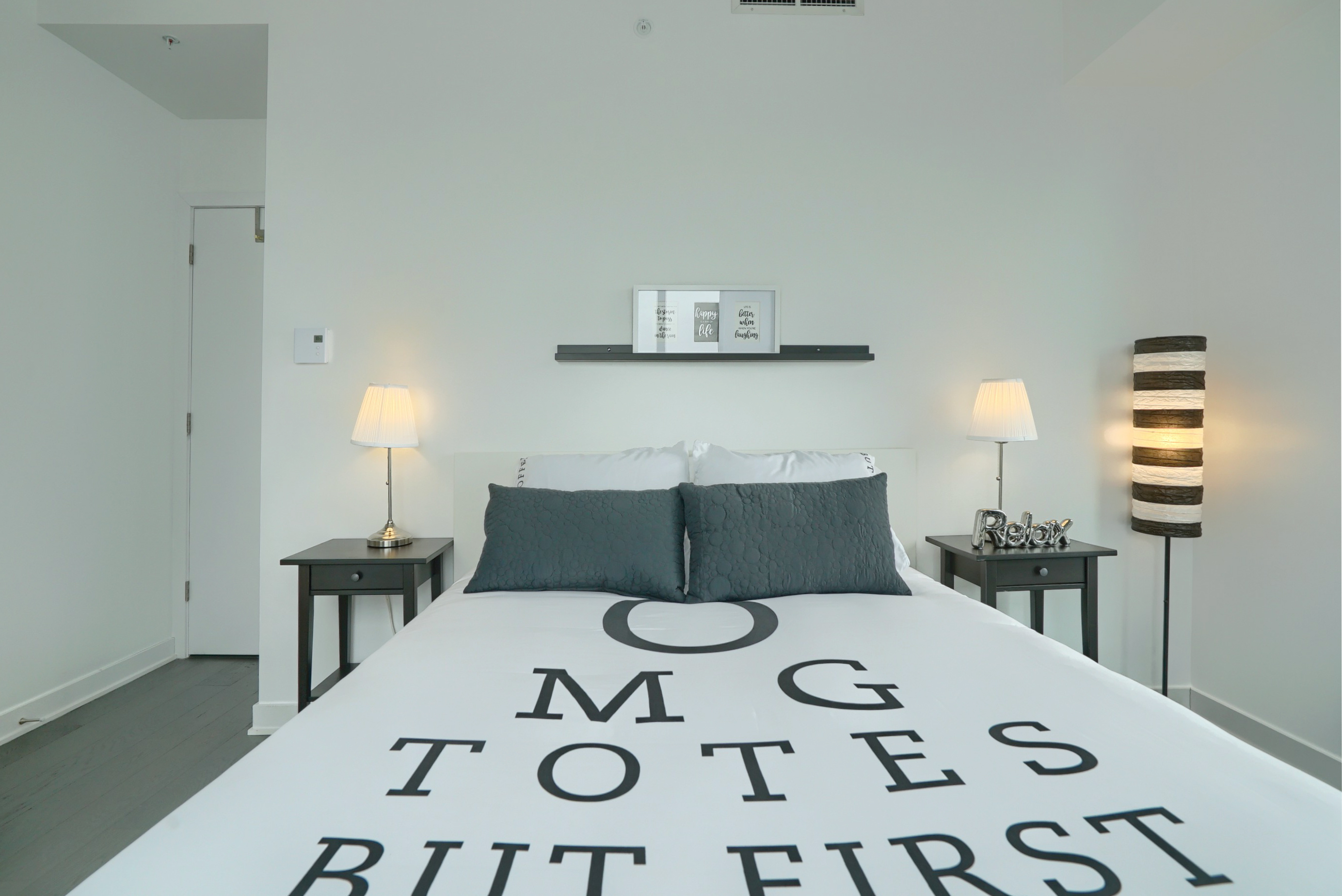 Gros plan du lit du pied du lit au mur. Couvre-lit unique - blanc avec des lettres grises centrées. Des lampes design et des supports de lit de chaque côté en font un loft meublé centre-ville de Montréal 