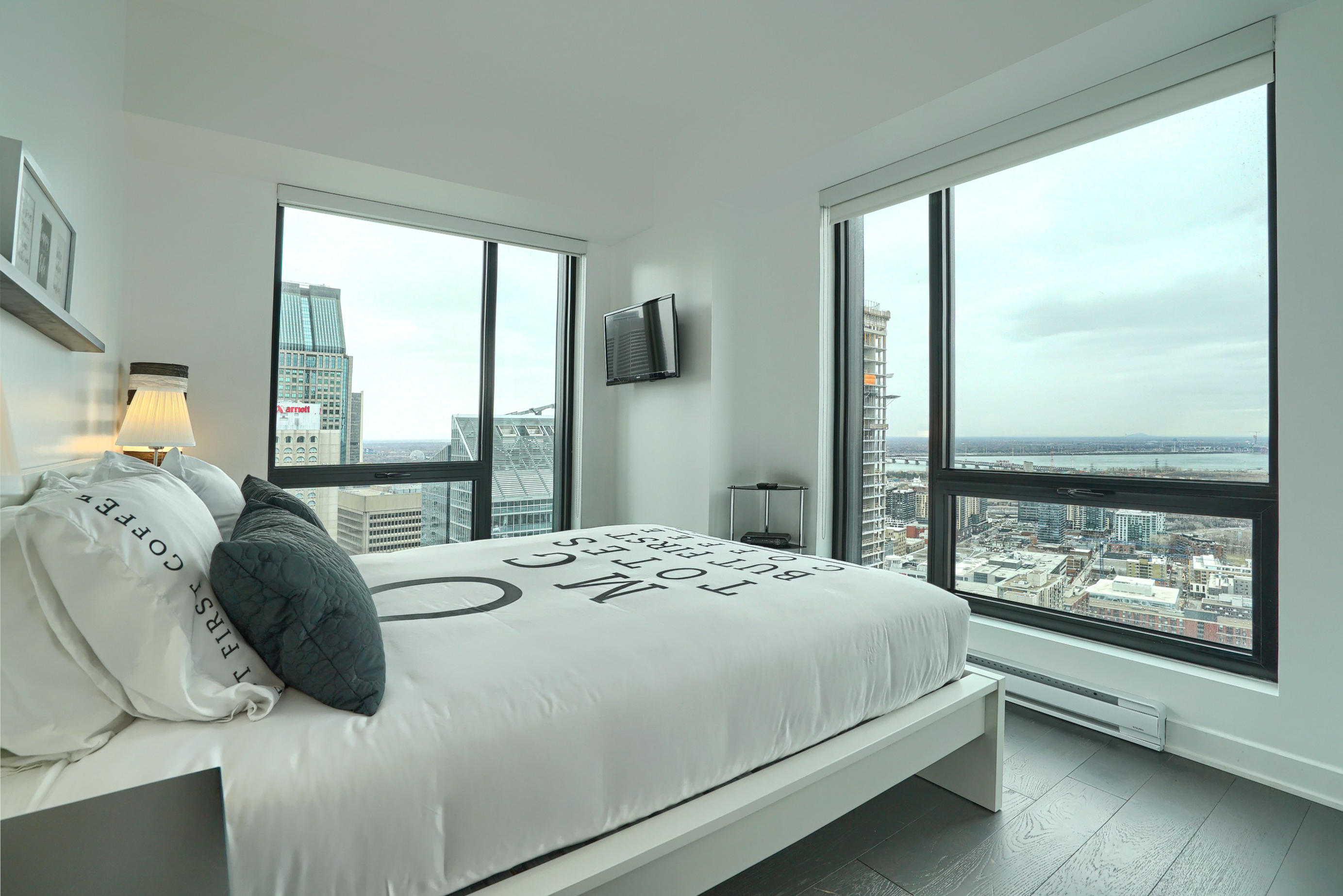 Vue en angle de la chambre montrant le côté du lit et les deux grandes fenêtres du sol au plafond pour rendre votre séjour dans ce condo meublé de luxe à Montréal 
unique
