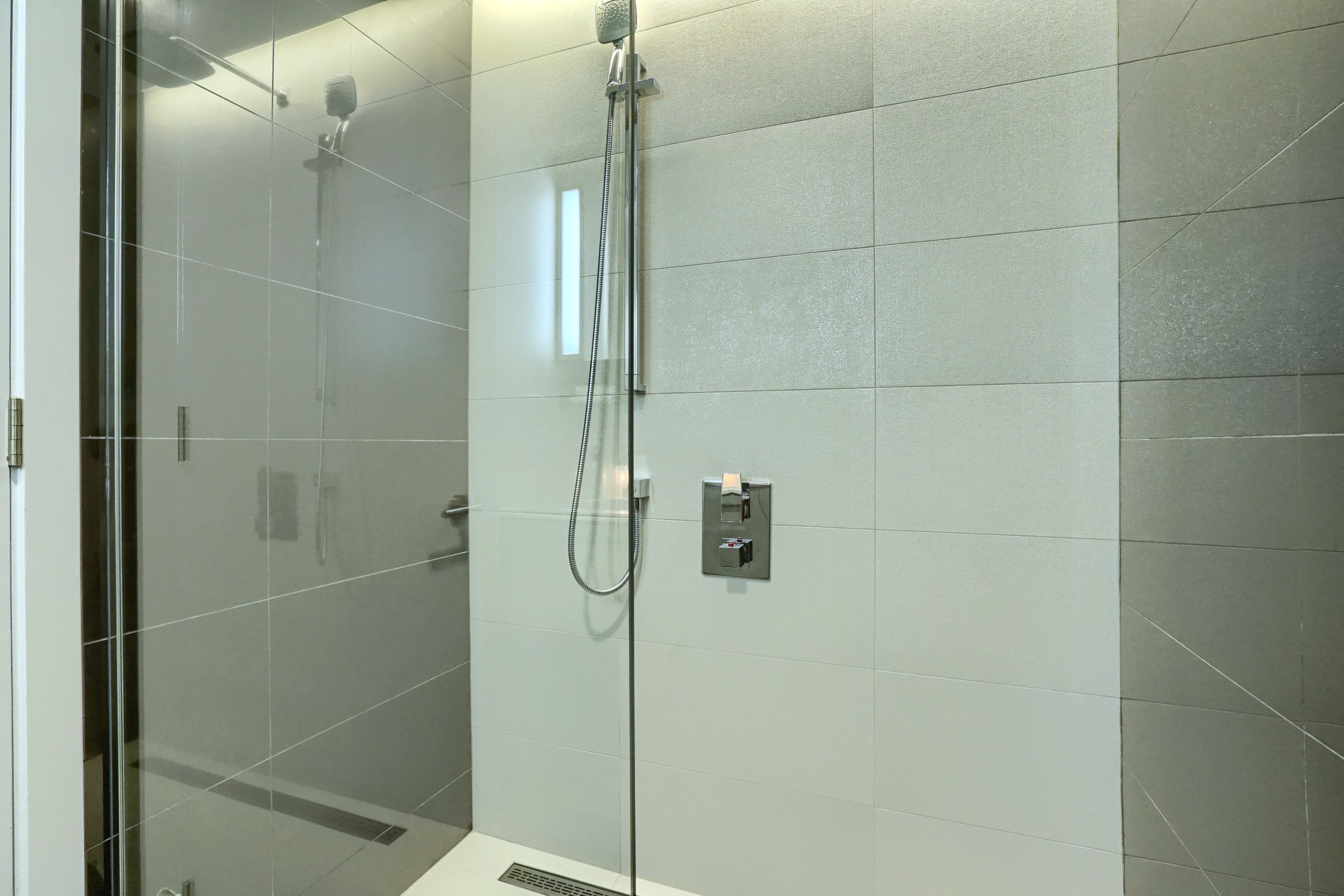 Vue rapprochée de la douche en verre du sol au plafond avec pomme de douche réglable. Accents inoxydables avec carreaux élégants blancs et gris. Salle de bain super moderne dans cet logement meublé à Montréal 